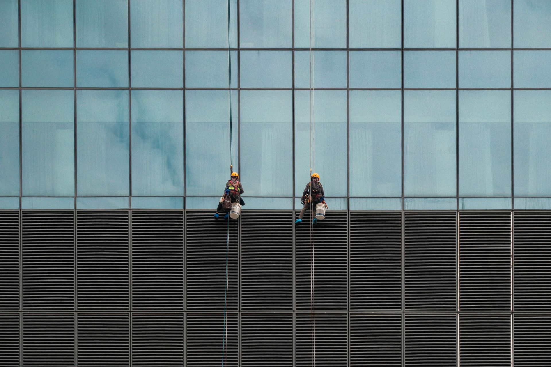 Two window cleaners suspended on ropes cleaning a modern skyscraper facade.